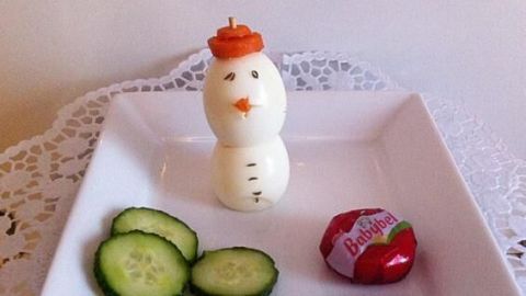 Ein Schneemann aus Eiern steht auf einem weißen Teller, umgeben von Gurkenscheiben und einer Süßigkeit.
