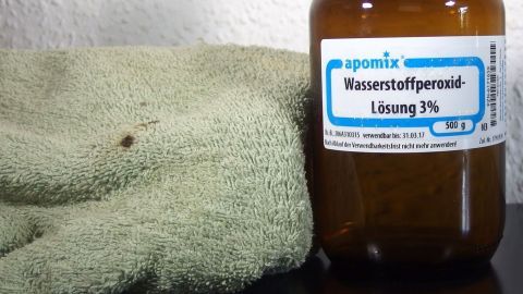 Wasserstoffperoxid hat ein Sauerstoffteilchen mehr als Wasser, von daher löst es das Blut leichter/besser. Ein Fläschchen mit 3% Wasserstoffperoxid steht neben einem grünen Tuch, um Blutflecken zu entfernen.
