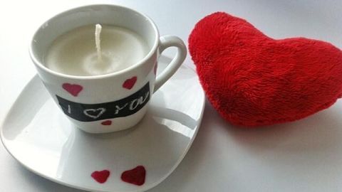 Eine Kerze in einer Tasse mit Herzmotiven steht auf einer Untertasse, während daneben ein rotes Herz aus Wolle liegt.