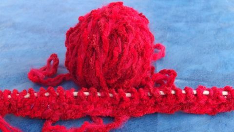 Ein rotes Wollknäuel liegt auf einer blauen Unterlage und wird verwendet, um eine Strickarbeit anzufertigen.