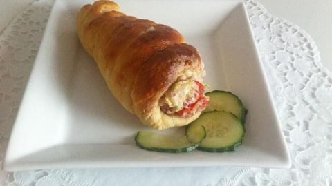Eine gefüllte Brötchenrolle mit Thunfischsalat liegt auf einem weißen Teller, umgeben von Gurkenscheiben auf einem Tisch.