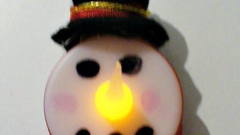 Ein LED-Schneemann in Teelichtoptik leuchtet, mit Hut und Schal geschmückt, an einer Schnur hängend vor weißem Hintergrund.