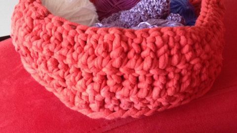 Ein roter Korb aus Jersey-Betttuch liegt auf einem Sofa und enthält bunte Wollen und Knäuel in verschiedenen Farben.