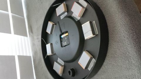 Eine runde, schwarze Uhr mit vielen kleinen, quadratischen Ziffern zeigt die Zeit an. Sie liegt auf einer grauen Oberfläche.