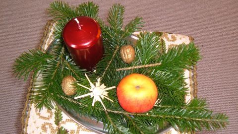 Ein Adventsgesteck mit einer roten Kerze und einem Apfel, umgeben von Tannenzweigen, auf einem dekorativen Tisch.