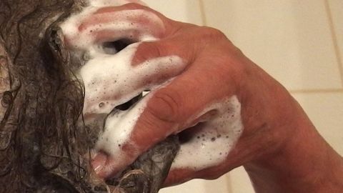 Eine Hand mit Seife reinigt schonend die Oberfläche eines Ohres. Der Hintergrund ist eine Badezimmerumgebung.