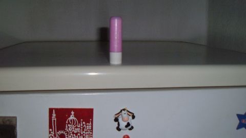 Auf der Oberseite eines Kühlschranks steht ein rosa Lippenstift, umgeben von Kühlschrankmagneten und einer grauen Wand.