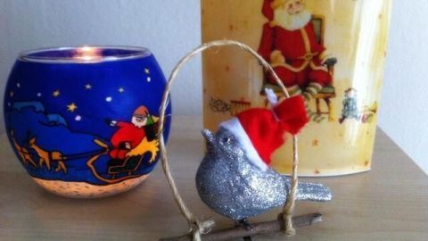 Ein glitzernder Weihnachtsvogel mit rotem Hut hängt an einem Zweig, umgeben von einem dekorativen Teelichthalter und einer Weihnachtskerze.