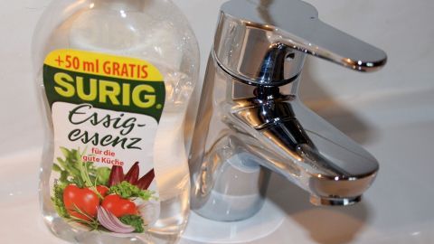 Eine Flasche Essigessenz steht neben einem glänzenden Wasserhahn und verdeutlicht die Anwendung säurehaltiger Entkalker.