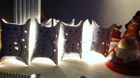 Eine Lichterkette mit Schneemann-Dekoration leuchtet sanft auf einer festlich geschmückten Oberfläche, umgeben von Weihnachtsfiguren.