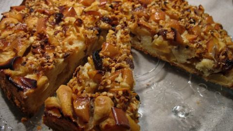 Eine Apfeltorte mit knuspriger Nuss- oder Mandelkruste liegt auf einem durchsichtigen Teller und hat ein Stück herausgeschnitten.