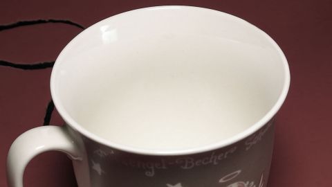 Eine weiße Tasse mit Engelsmotiv steht auf einem roten Hintergrund. Rundum ist der Schriftzug "Engel-Becher" in Weiß zu sehen.