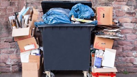 Eine überfüllte Mülltonne mit blauen Säcken ist von Kartons und Papier umgeben, vor einer Steinmauer platziert.