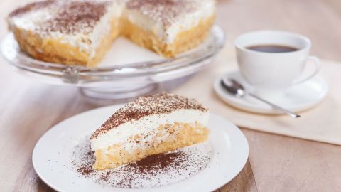 Eine schwedische Apfeltorte liegt auf einem Teller, während ein Stück abgeschnitten ist, um den cremigen Belag zu zeigen. Im Hintergrund steht eine Tasse Kaffee.