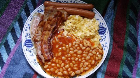 Ein Teller mit englischem Frühstück, der gebratenen Speck, Würstchen, Rührei und gebackene Bohnen präsentiert, auf einem bunten Tuch.