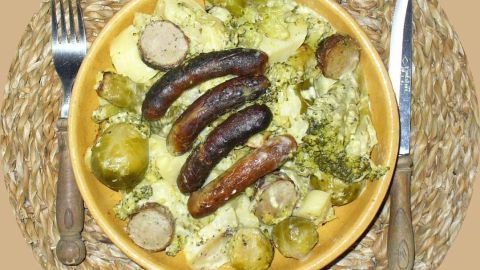 Ein Gemüseauflauf ist in einer Schüssel zu sehen, auf dem mehrere Grillbratwürste liegen, umgeben von verschiedenen Gemüse.