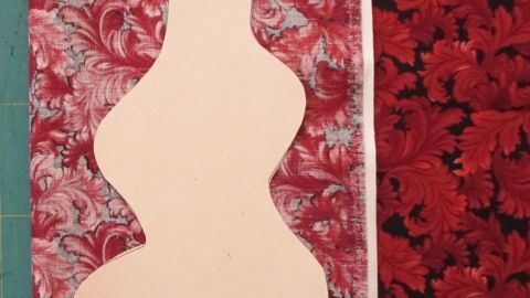 Ein stofflicher Tannenbaum-Schnitt sitzt auf rotem, gemustertem Stoff in einer bastelnden Umgebung, um kreativ gestaltet zu werden.