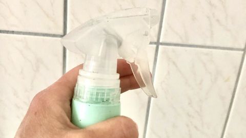 Eine Hand hält eine Sprühflasche, während in der Dusche die Fliesen mit Schimmelflecken im Hintergrund sichtbar sind.