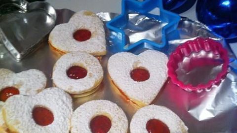 Spitzbuben-Kekse mit Puderzucker und Marmelade liegen auf Alufolie, umgeben von Ausstechformen und blauen Kugeln.