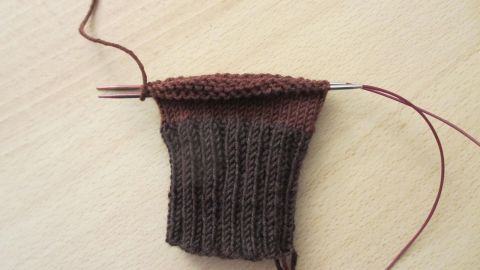 Eine gestrickte Socke mit einem dunkelbraunen, gerippten Bund liegt auf einer hellen Tischoberfläche, während der Magic Loop Technik verwendet wird.