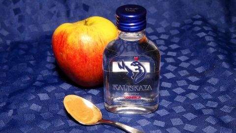 Ein roter Apfel liegt neben einer kleinen Flasche klaren Likörs mit einem Löffel auf einer blauen Unterlage.