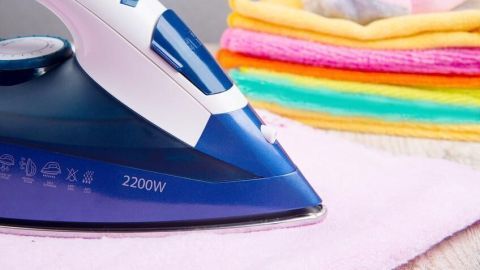 Ein blau-weißes Bügeleisen mit der Aufschrift "2200W" gleitet über ein rosa Tuch, daneben bunte Handtücher.