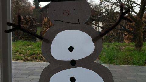 Ein Schneemann aus Papier ist am Fenster befestigt, mit einem schwarzen Hut, Arme aus Zweigen und fröhlichem Gesicht.