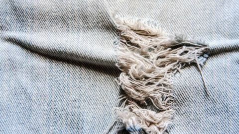 Eine zerrissene Kinderhose zeigt ausgefranste Fasern auf blauem Stoff, die geflickt werden müssen.