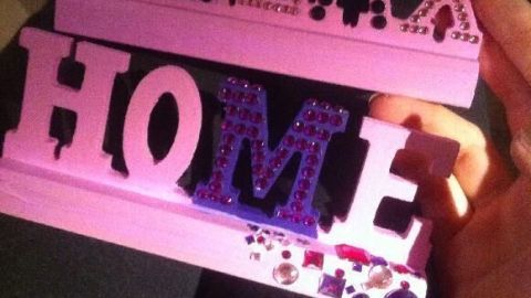 Eine Hand hält ein verziertes, rosa Schild. Darauf steht "RELAX HOME" mit glitzernden Steinchen verziert, auf schwarzem Stoff.