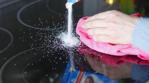 Eine Hand streut Reinigungspulver auf eine Herdplatte, während mit einem Tuch hartnäckige Verkrustungen entfernt werden.