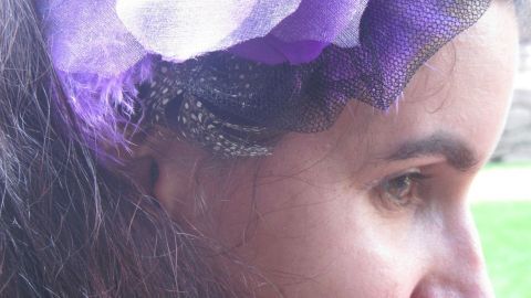 Ein eleganter Fascinator mit lila Blumen ziert das Haar einer Frau, während sie in einer grünen Umgebung steht.