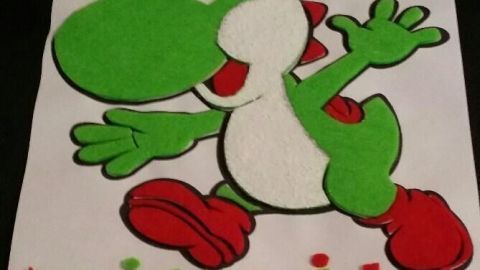 Ein grüner 3D-Yoshi aus Filz springt fröhlich auf einem weißen Hintergrund. Darunter steht der Name "Mikail" in bunten Buchstaben.
