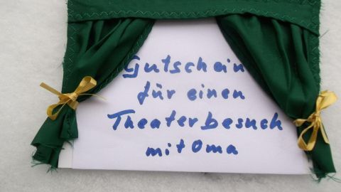 Ein Gutschein für einen Theaterbesuch mit Oma liegt dekorativ hinter einem Vorhang aus grünem Stoff.