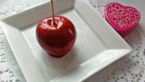 Ein karamellisierter Apfel steht auf einem weißen Teller mit Zuckerguss, umgeben von einer herzförmigen Dose.