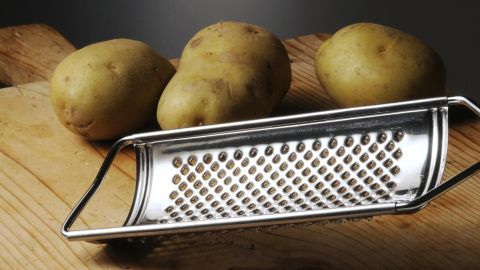 Eine Reibe liegt auf einem Holztisch neben drei Kartoffeln, bereit zum Raspeln.