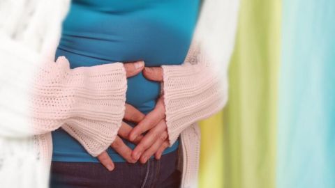 Hände halten einen schmerzenden Bauch, während eine Person in einem blauen Shirt und weißer Strickjacke im bunten Raum steht.