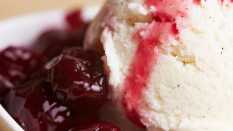 Eine Kugel Vanilleeis liegt in einer Schüssel, überzogen mit heißen Kirschen und roter Soße, umgeben von einem Holzuntergrund.