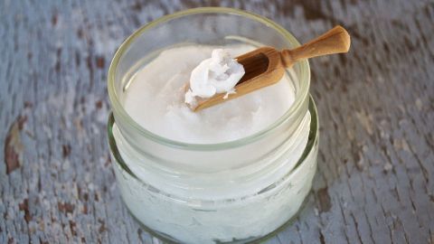 Ein Glasbehälter mit Kokosöl, ein Holzlöffel entnimmt das Öl aus dem Behälter auf einer rustikalen Oberfläche.