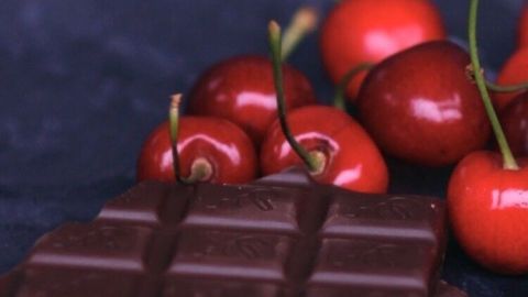 Ein Stück Schokolade liegt neben frischen Kirschen auf einem dunklen Untergrund, was einen köstlichen Dessertkontext schafft.