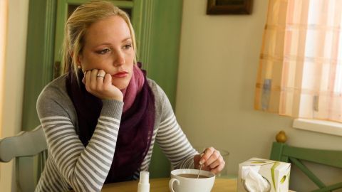 Eine Frau sitzt nachdenklich an einem Tisch, trinkt Tee und sieht müde aus, umgeben von Erkältungsmedikamenten.