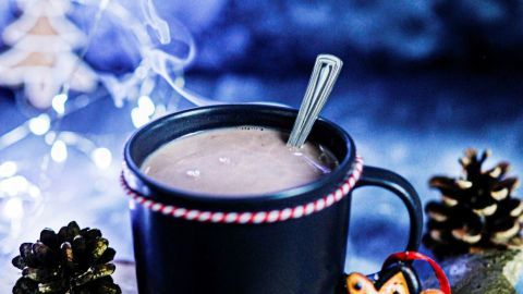 Schwarze Tasse mit dampfendem Kakao steht auf einem Holzbrett, umgeben von Weihnachtsdekoration und Gewürzen.