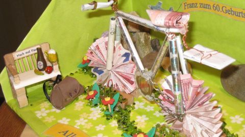 Ein aus Geldscheinen gefaltetes Fahrrad steht auf einer grünen Unterlage mit Blumen und einer kleinen Bank.