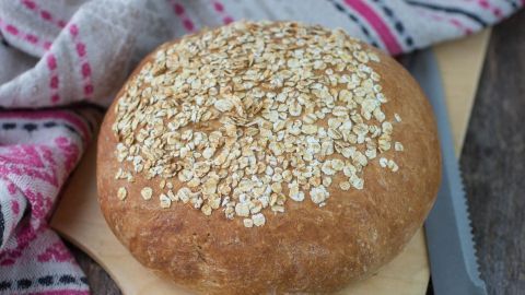 Rundes Haferflocken-Quark-Brot liegt auf einem Holzbrett, umgeben von einem bunten Tuch und einem Messer.