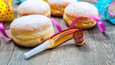 Fasnachtsküchle mit Puderzucker liegen auf einem Holztisch, umgeben von bunten Luftschlangen und einer Partytröte.