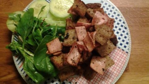 Ein Teller mit gewürfeltem Pausenbrot, belegt mit Wurst und serviert mit Gurkenscheiben und frischem Salat auf einem Holztisch.