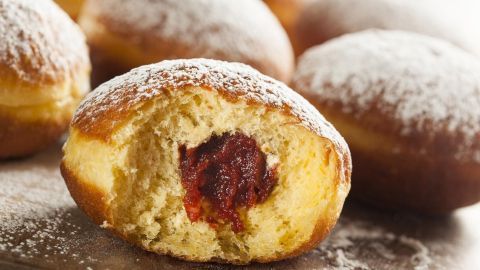 Ein Berliner Pfannkuchen liegt auf einem Holzbrett. Er ist halbiert, zeigt die süße Füllung und ist mit Puderzucker bestäubt.