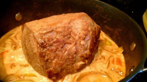 Ein saftiger Rinderbraten liegt in einer tiefen Pfanne in reichhaltiger Soße, umgeben von Zwiebeln und Karotten.