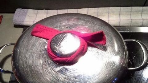 Ein heißer Kochtopfdeckel liegt auf einem Herd, mit einem pinken Tuch als Schutz vor der Hitze im Küchenumfeld.