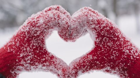 Rote Schneehandschuhe formen ein Herz, während sie von einer dünnen Schicht Schnee bedeckt sind, umgeben von Winterlandschaft.