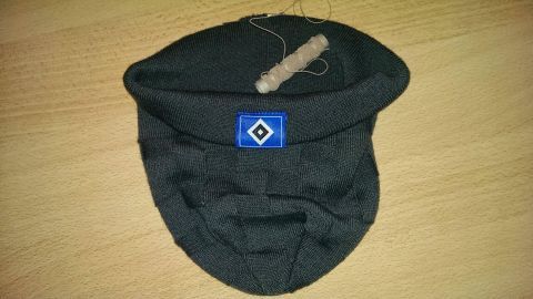Ein schwarzer Mützenrand sitzt auf einem Tisch, während ein Stück Hutgummi daneben liegt, bereit zur Befestigung.